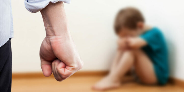 Family violence and aggression concept - furious angry man raised punishment fist over scared or terrified child boy sitting at wall corner